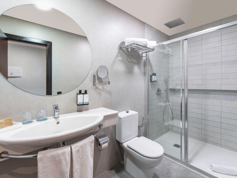 Moderner Badezimmer mit ovalem Spiegel, weißem Waschbecken, Toilette und Glaskabine für Dusche.