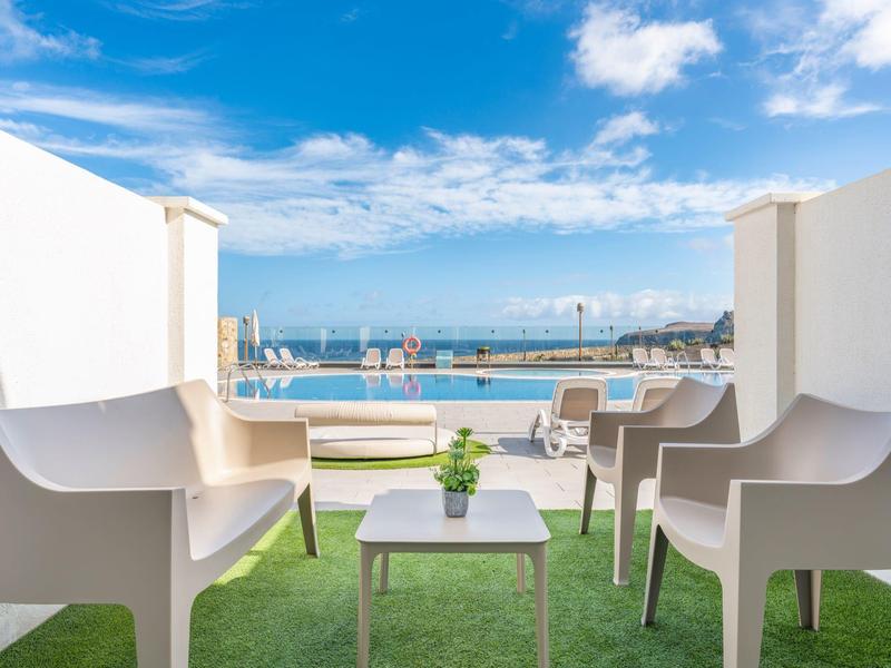 Balkon mit weißem Mobiliar, grünem Kunstgras und Blick auf Pool und Meer unter blauem Himmel.