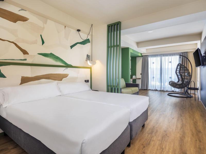 Modernes Hotelzimmer mit zwei weißen Betten, Holzboden, grünem Akzent und einem hängenden Schaukelstuhl.