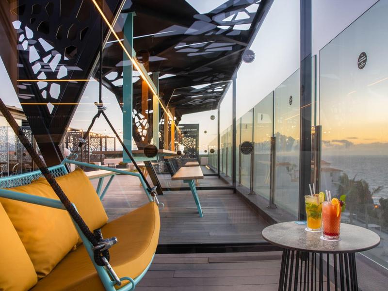 Moderner Balkon mit gelben Hängesesseln, Glastür, Holzterrasse und zwei Getränken auf kleinem Tisch.