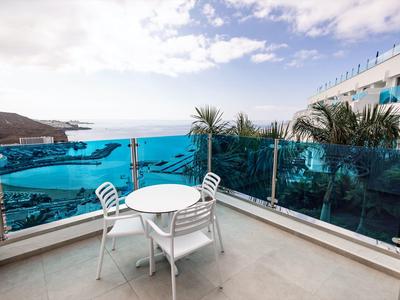Balkon mit weißem Tisch und Stühlen, blauer Glasbrüstung, Blick aufs Meer und Himmel mit Wolken.