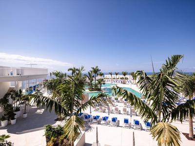 Hotel moderno con piscina, palme e lettini sotto un cielo blu limpido.