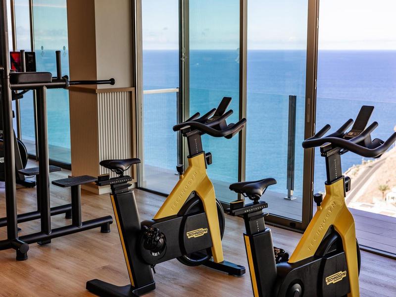 Due cyclette gialle in una palestra con grandi finestre e vista sul mare.
