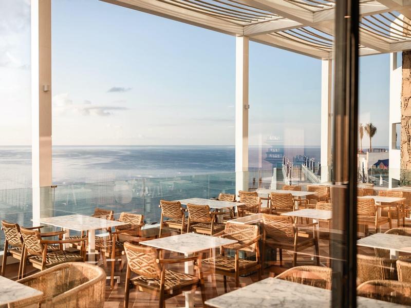 Terrazza soleggiata con tavoli e sedie di legno con vista sul mare.
