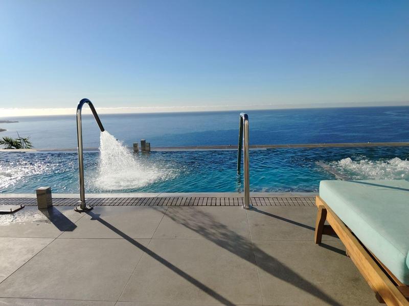 Una piscina a sfioro con cascata e vista sul mare, lettino con cuscino blu al bordo.
