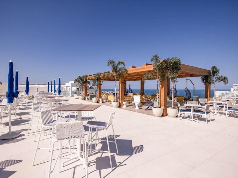 Terrazza panoramica con tavoli bianchi, sedie e pergolato in travi di legno con vista sul mare