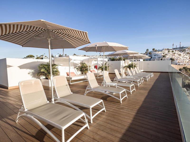 Terrazza soleggiata con lettini e ombrelloni sul tetto di un hotel sotto un cielo limpido.