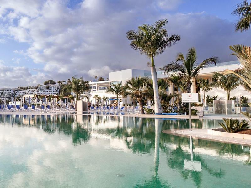 Piscina dell'hotel moderna con palme sotto un cielo azzurro e edifici bianchi sullo sfondo.