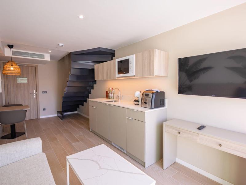 Camera d'hotel luminosa con angolo cottura moderno, scala e grande TV a muro.