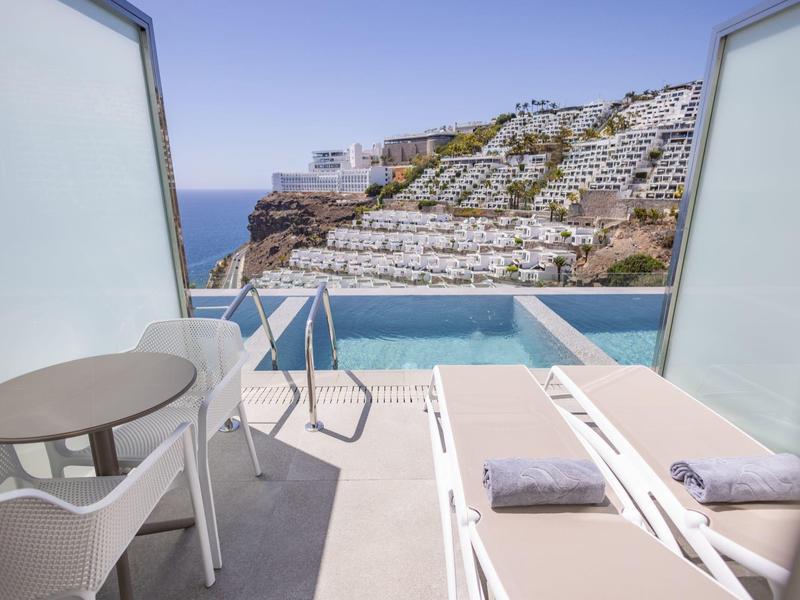 Balcone con piscina, due lettini e vista mare in un hotel soleggiato.