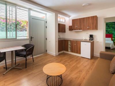 Helles Wohnzimmer mit Holzboden, Sofa, kleinem Tisch, Küchenzeile und zwei Stühlen am Fenster.