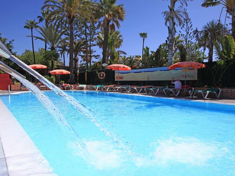 Buitenzwembad met waterstralen, omringd door palmbomen en ligstoelen met oranje parasols.