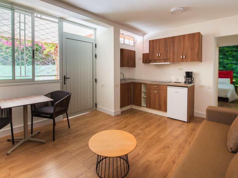 Lichte hotelkamer met houten vloer, kitchenette, zithoek en raam met gordijnen.