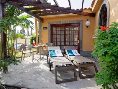Dos tumbonas y una mesa frente a una habitación de hotel amarilla con plantas y pérgola.