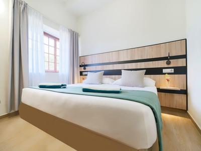 Chambre d'hôtel moderne avec grand lit, parquet en bois et fenêtre avec rideaux blancs.