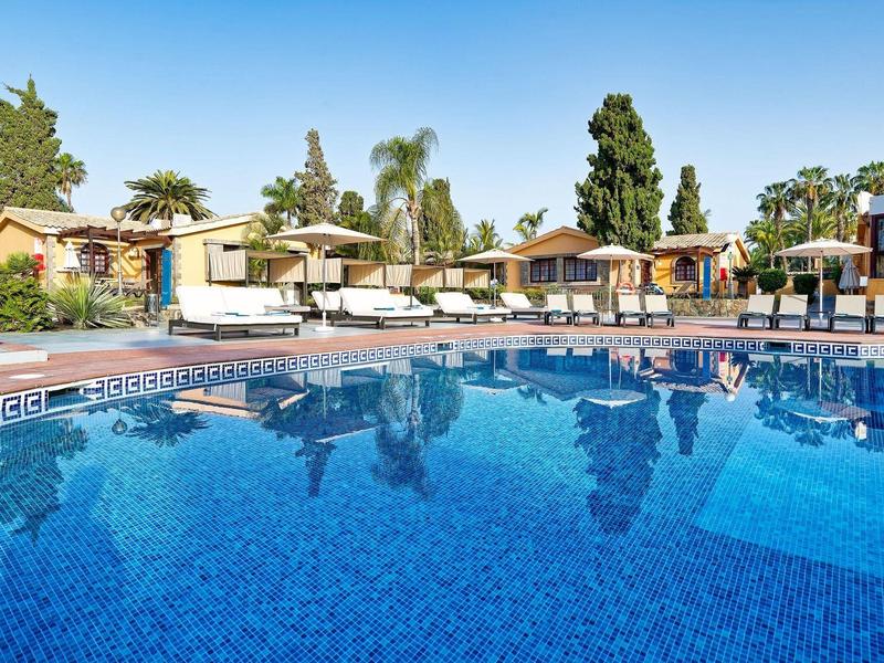 Complexe hôtelier avec piscine claire, parasols et bungalows en arrière-plan sous un ciel bleu.