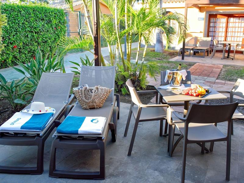 Terrasse avec chaises longues et table avec fruits dans un jardin ensoleillé.