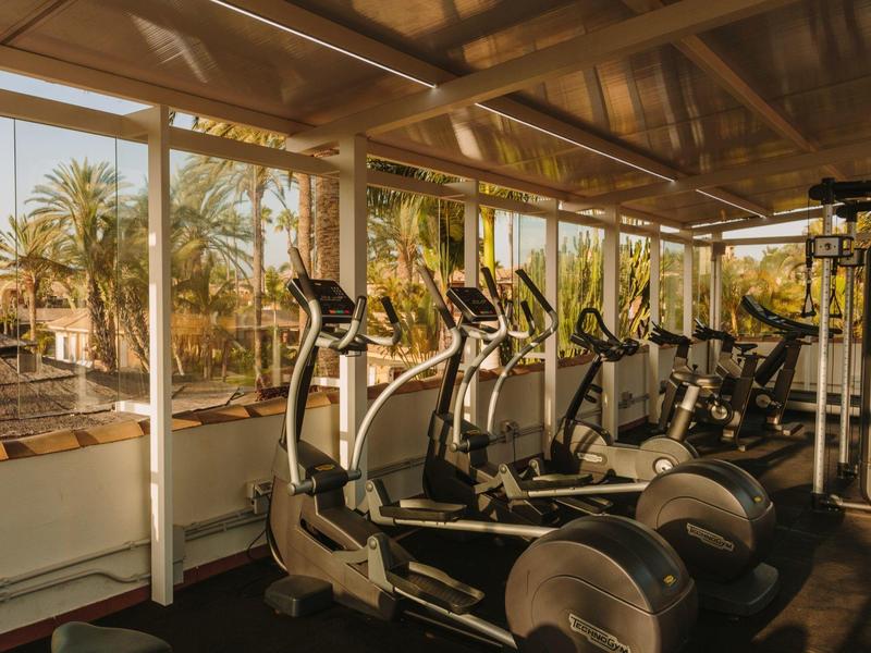 Salle de sport avec plusieurs tapis de course et elliptique, entourée de grandes fenêtres donnant sur des palmiers.