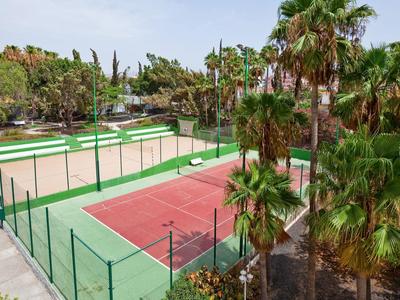 Campo da tennis rosso con recinzione verde circondato da palme in una zona soleggiata.