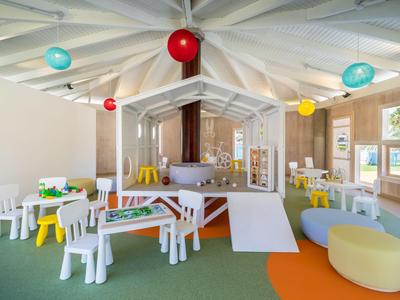 Area gioco per bambini colorata con tavoli, sedie e uno scivolo piccolo in una stanza luminosa.