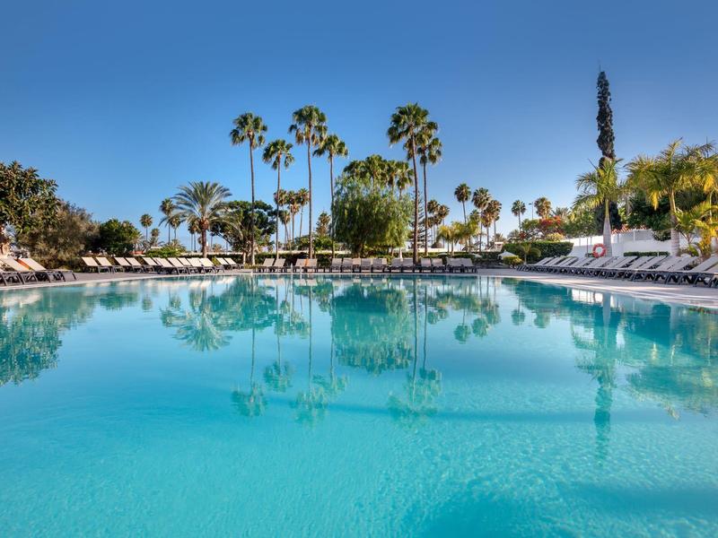 Piscina de hotel con agua azul y palmeras bajo un cielo despejado con tumbonas.