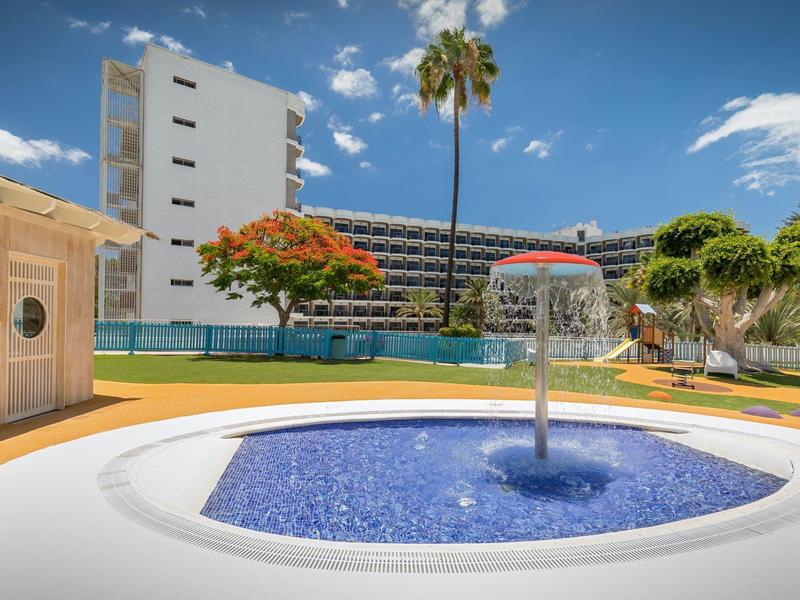 Piscina moderna de hotel con fuente de agua, rodeada de árboles y edificios de varios pisos bajo cielo azul.