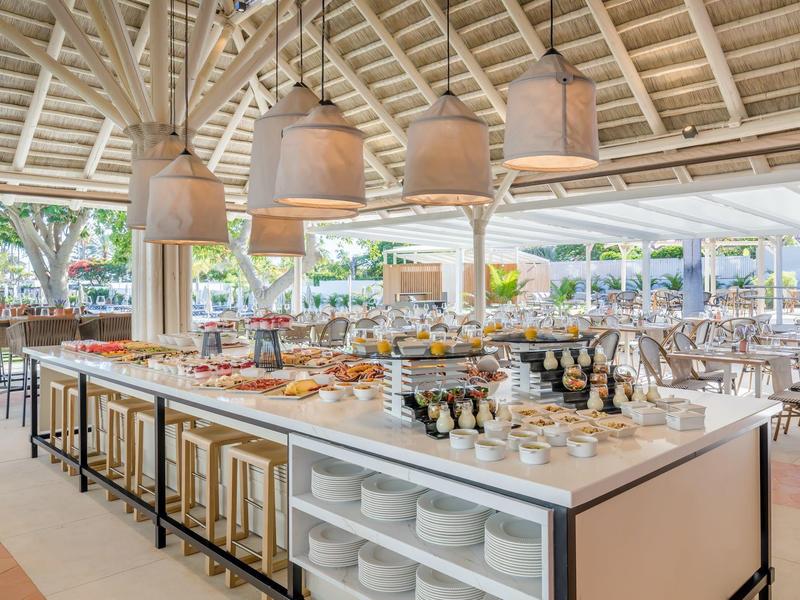 Buffet de restaurante abierto con taburetes, lámparas y mesas preparadas bajo un techo de madera.