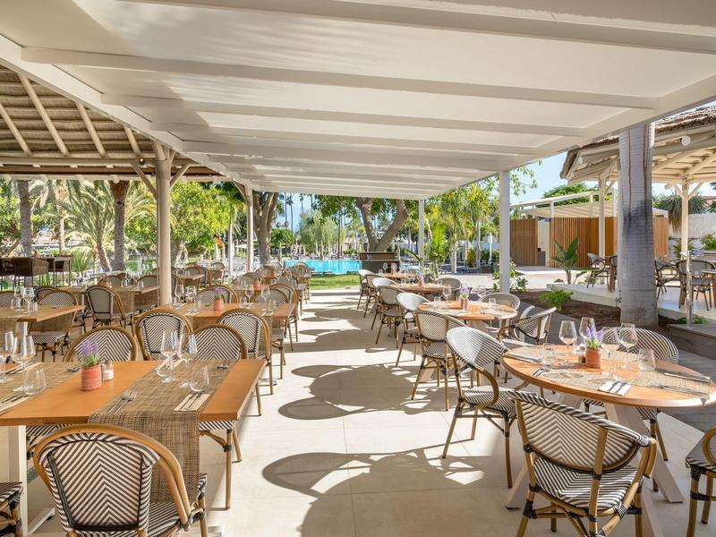 Área externa abierta de un restaurante con sillas de mimbre y mesas de madera bajo un toldo, palmeras al fondo.