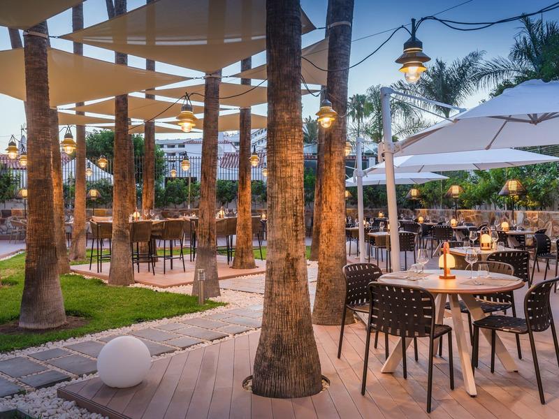 Acogedora zona exterior de restaurante con mesas, sillas y palmeras al atardecer.