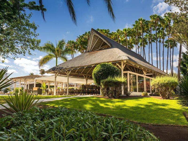 Edificio de hotel tropical con techo de palma rodeado de palmeras verdes y césped bajo cielo despejado.