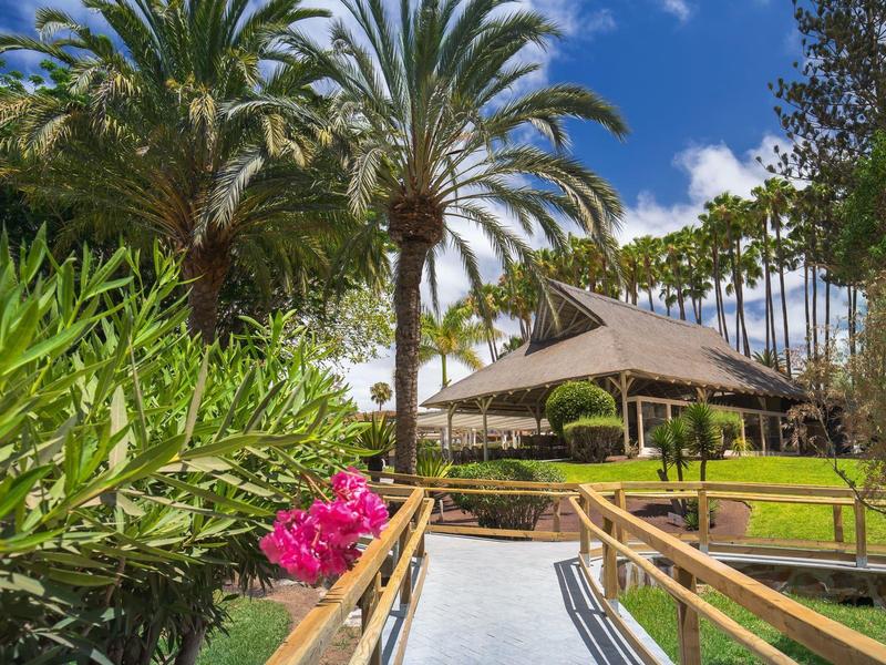 Camino de madera rodeado de vegetación florida que conduce a una cabaña en un entorno tropical soleado.