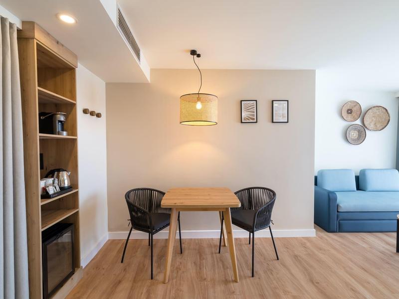 Área de comedor moderna con mesa de madera, dos sillas y sofá azul en habitación luminosa con suelo de madera.