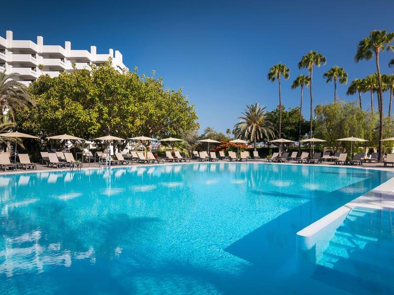 Gran piscina con tumbonas y palmeras frente a un hotel de varios pisos bajo un cielo despejado.