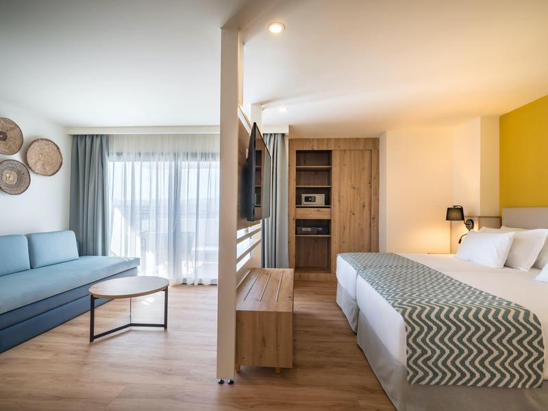 Habitación de hotel moderna con cama doble, sofá, muebles de madera y grandes ventanas con cortinas.