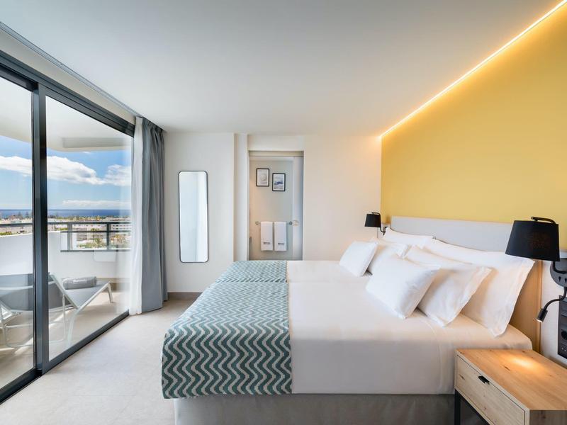Habitación moderna de hotel con cama doble, pared amarilla y balcón con vista al mar.