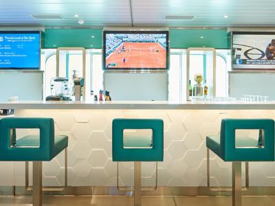 Area bar moderna in hotel con banconi bianchi e sgabelli turchesi, illuminata e con schermi.