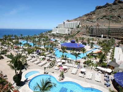 Large hotel complex with multiple pools, sun loungers, and sea view in a mountainous setting.