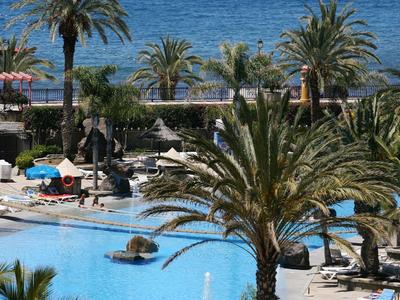 Large outdoor pool with loungers, palm trees, and a view of the blue sea in sunny weather.