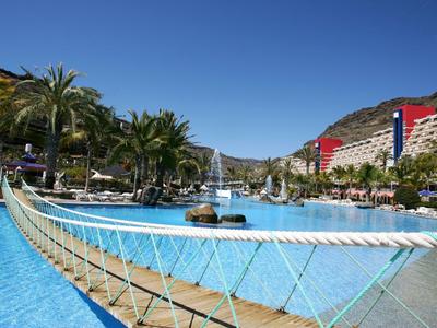 Large hotel pool with suspension bridge, palm trees, and hotel buildings under clear sky.