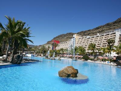 Large outdoor pool with rocks and palm trees in front of a hillside hotel under clear sky.