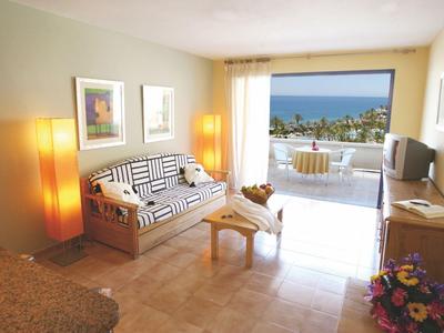 Living room with sofa, armchair, glass table, and balcony with sea view in a hotel room.