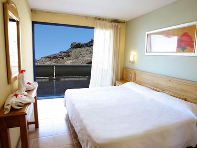 Bright hotel room with large bed and balcony overlooking a mountain.