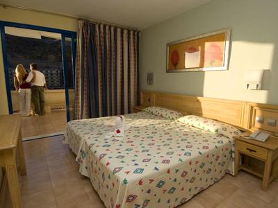 Bright hotel room with double bed, nightstands, and a couple on the balcony at sunset.