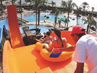 Two children slide down an orange water slide in a tropical pool area with palm trees.