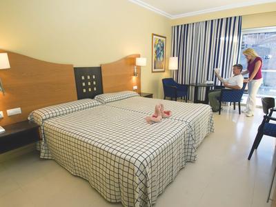 Bright hotel room with two single beds and guests near window overlooking city.