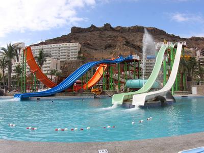 Several water slides lead into a large pool with rocky cliffs and hotel buildings in the background.