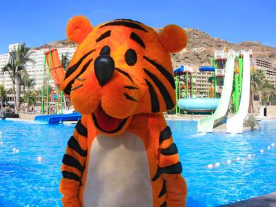A tiger mascot stands in front of a pool with water slides and palm trees under clear skies.