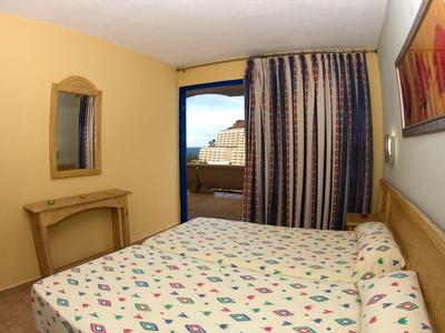 Small hotel room with colorful bedspread, wooden furniture, and balcony overlooking the sea.