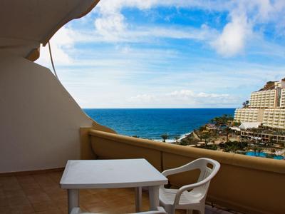 Balcony with table and chair overlooking sea and hotel buildings under blue sky.