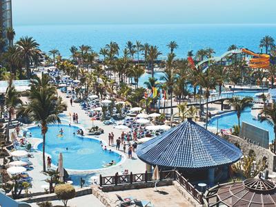 Großer Hotelpool mit Palmen und Gästen, dahinter Blick auf das blaue Meer und den Himmel.